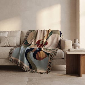 May include: A woven throw blanket featuring an abstract design with curved shapes in shades of light blue, dark navy, deep red, olive green, and cream. The blanket has a textured surface and decorative fringe along the edges, draped over a sofa.