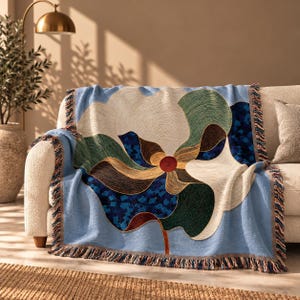 May include: A woven throw blanket featuring a large, abstract floral design in shades of cream, dark green, brown, and blue. The blanket has a light blue background, a central red circular accent, and decorative fringe along the edges.