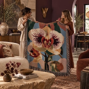 Puede incluir: Una manta tejida que presenta un diseño grande y detallado de flor de orquídea en tonos crema, amarillo y burdeos intenso sobre un fondo azul claro, acabado con un borde decorativo de flecos en todos los lados del tejido rectangular.