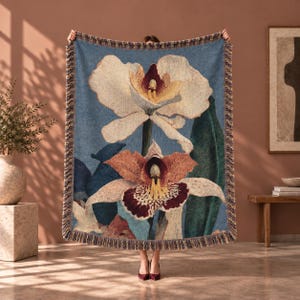 May include: A woven throw blanket featuring a large, detailed botanical print of two orchids against a blue background. The top orchid is cream and yellow, while the bottom orchid displays orange, maroon, and yellow petals. The edges have a fringe.