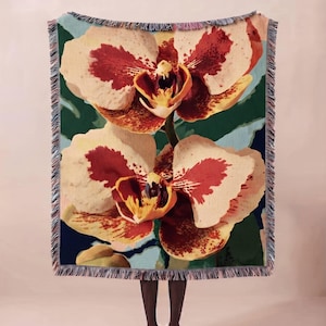 May include: A rectangular woven throw blanket featuring a large-scale botanical print of two cream and deep red orchids against a dark green foliage background. The blanket has a decorative fringed edge with multi-coloured threads on all four sides.