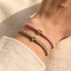 May include: Two macrame bracelets, one pink and one tan, are worn on a wrist. Each bracelet has a gold heart charm with a letter. The pink bracelet has a "J" charm, and the tan bracelet has an "A" charm. The bracelets are on a light-colored background.