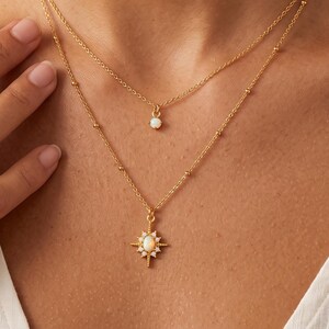 Puede incluir: Un conjunto de collares dorados en capas con dos cadenas delicadas. La cadena más corta tiene un pequeño colgante de piedra blanca redonda, mientras que la más larga incluye un colgante en forma de estrella con una piedra blanca central.