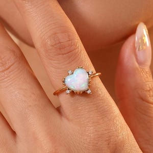 May include: A gold ring featuring a heart-shaped opal gemstone as the centerpiece, accented by small, clear, round-cut stones set into the metal prongs surrounding the opal. The band is thin and polished, showcasing a simple and elegant design.