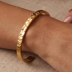 May include: A gold-toned metal cuff bracelet featuring a textured, hammered surface finish. The bracelet has a C-shaped open design that wraps around the wrist, showcasing a reflective, metallic sheen across its entire outer circumference.