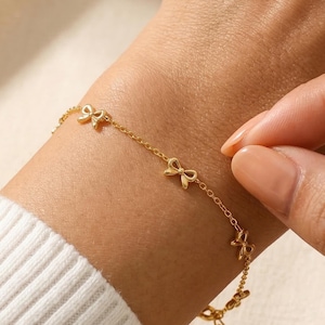 Puede incluir: Una pulsera dorada con una delicada cadena y varios dijes en forma de lazo. La pulsera se lleva en la muñeca, con el cierre ajustable visible. La joya tiene un cálido tono dorado y el fondo es de un color suave y neutro.