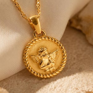 Può includere: Una collana color oro con un ciondolo circolare caratterizzato da un rilievo di un cherubino alato. Il ciondolo è incorniciato da un bordo a corda intrecciata e pende da una sottile e scintillante catena color oro con maglie intrecciate.