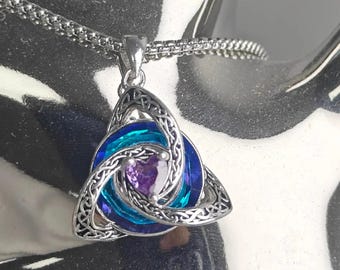 May include: A silver-toned pendant necklace featuring a triquetra-inspired knotwork frame surrounding a circular blue and teal crystal element with a small, central purple heart-shaped stone, suspended from a matching silver-toned chain.