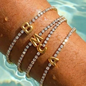 Op de afbeelding: Vier goudkleurige tennisarmbanden met diamantachtige accenten en initiaalbedels die de letters B, M, C en A spellen. De armbanden worden om een pols gedragen, gedeeltelijk ondergedompeld in water, met een blauwe achtergrond.