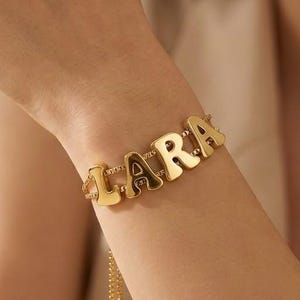 Pode incluir: Uma pulseira dourada com o nome "LARA" escrito em letras de bloco individuais. A pulseira é usada no pulso e tem uma corrente delicada para ajustar o tamanho. As letras são contornadas em dourado com um preenchimento mais escuro.