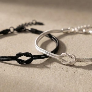 May include: Two bracelets, one black and one silver, each featuring a knot design. The bracelets are on a neutral-toned surface, with the black bracelet on the left and the silver one on the right. The bracelets have adjustable clasps.