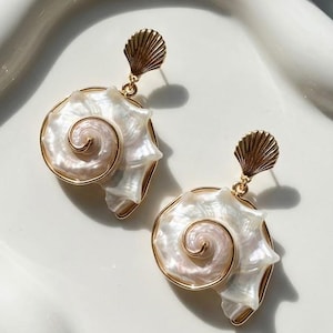 May include: A pair of gold-toned earrings featuring iridescent white spiral seashell charms. Each earring has a small, fan-shaped gold metal topper attached to a polished shell component that displays a natural pearlescent sheen and spiral design.