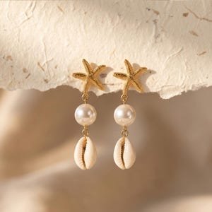 May include: Gold-coloured starfish earrings with a pearl and seashell drop. The earrings feature a textured starfish design at the top, a large white pearl in the middle, and a small, cream-coloured seashell at the bottom. The earrings are designed for pierced ears.