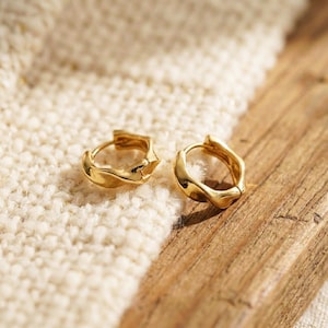 Può includere: Un paio di piccoli orecchini a cerchio color oro. Gli orecchini hanno un design strutturato e organico. Sono esposti su una superficie bianca strutturata e una superficie in legno.