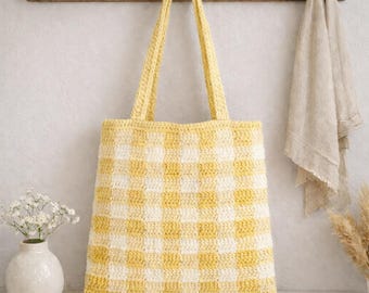 May include: A handmade crochet tote bag in a yellow and white checkered pattern. The bag has two long handles and is hanging on a wooden rack. The bag is made of a soft, textured yarn and is perfect for carrying groceries or other items.