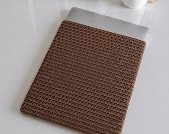 May include: A brown, crocheted laptop sleeve. The sleeve is made of a textured, ribbed material and is designed to fit a laptop. The laptop is partially visible, with the top portion of the device exposed. A white mug with a gold handle and a small white vase are in the background.