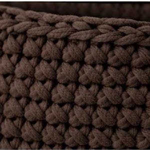 May include: Close-up of a brown, chunky knit textile. The fabric is made of thick, intertwined loops, creating a textured surface. The color is a deep, earthy brown, and the knit pattern is consistent throughout the image.