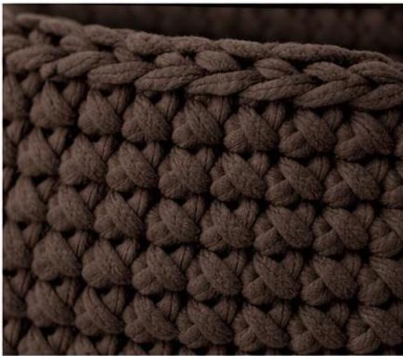 May include: Close-up of a brown, chunky knit textile. The fabric is made of thick, intertwined loops, creating a textured surface. The color is a deep, earthy brown, and the knit pattern is consistent throughout the image.