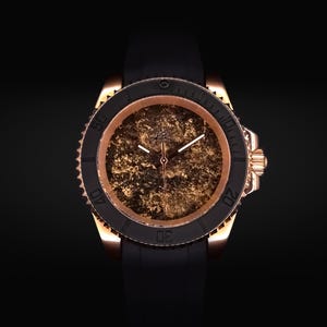 Puede incluir: Un reloj de pulsera de oro rosa con bisel negro y correa negra. La esfera del reloj tiene un diseño dorado texturizado. El reloj tiene una corona en el lado derecho. El fondo es negro.