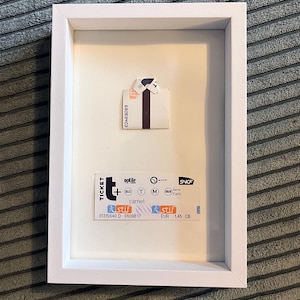 Può includere: Opera d'arte incorniciata con una camicia di carta piegata e un biglietto del treno. Il biglietto mostra i loghi "optile carnet" e "STIF". L'opera è in una cornice bianca, su uno sfondo grigio strutturato. Il biglietto mostra anche "EUR 1,45 CB".