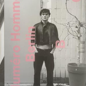 Può includere: Una copertina di rivista di moda in bianco e nero con una persona che indossa una giacca corta di pelle nera, pantaloni neri e stivali neri. Il titolo della rivista "Numéro Homme Berlin" è stampato in rosa.