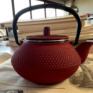 Puede incluir: Una tetera de hierro fundido roja con asa y boquilla negras. La tetera tiene una superficie texturizada y una tapa. La tetera está sobre una mesa con un libro abierto en primer plano.
