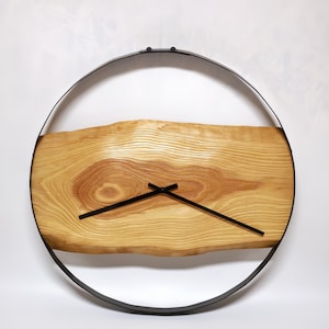 Könnte beinhalten: Eine runde Wanduhr mit einem hellbraunen Holzgesicht, das natürliche Maserungsmuster aufweist. Die Uhrzeiger sind schwarz, und das Zifferblatt ist in einem schwarzen Metallring eingefasst. Modernes, minimalistisches Design.