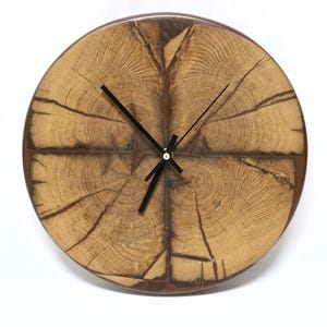 May include: Round wooden clock with a natural wood grain face. The clock features a dark brown frame and black clock hands. The face of the clock displays the natural patterns of the wood, creating a rustic aesthetic. The clock is a decorative item.