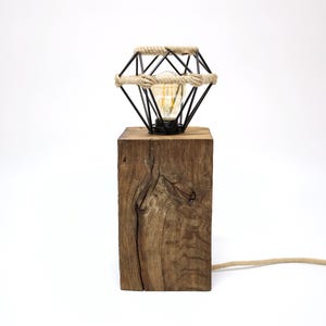Puede incluir: Lámpara de mesa rústica con pantalla de metal negro en forma de diamante, envuelta en cuerda, y una bombilla de estilo vintage. Se apoya sobre una base de madera rectangular y envejecida. El cable es de cuerda natural.