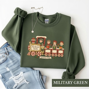 May include: A military green crewneck sweatshirt featuring a graphic of a steam train carrying four children holding gifts. The train engine is labeled The Brown Family and the banner below reads Christmas 2026. A silver necklace rests above.