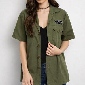 Op de afbeelding: Olijfgroen shirt met korte mouwen, een knoopsluiting, twee borstzakken en een kleine rechthoekige patch met de tekst "ROCK". Het shirt wordt gedragen over een zwarte top en gecombineerd met een spijkerbroek.