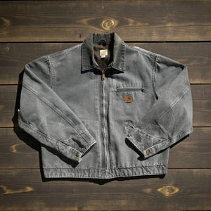 Può includere: Giacca da lavoro grigia con chiusura a zip completa, colletto e piccola tasca sul petto. La giacca ha una toppa con logo marrone e polsini con bottoni. La giacca è realizzata in materiale resistente.