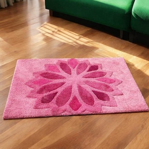 May include: A rectangular pink rug with a large, stylized flower design in shades of pink. The flower's petals are darker pink, creating a contrast against the lighter pink background. The rug is placed on a wooden floor.