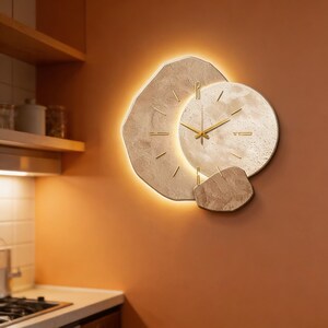 Puede incluir: Un reloj de pared moderno con un diseño único. El reloj presenta una esfera circular con manecillas y marcadores dorados sobre un fondo beige texturizado. El reloj está iluminado con una luz cálida y suave, creando un acento elegante.