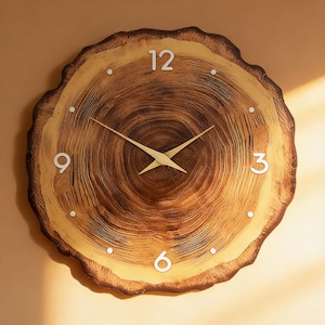 Op de afbeelding: Een ronde wandklok ontworpen om op een dwarsdoorsnede van een boomstam te lijken. De klok heeft een natuurlijke houtnerf met witte cijfers en goudkleurige wijzers. De klok heeft een diameter van ongeveer 30 cm.