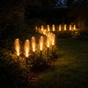 May include: A row of outdoor garden lights emitting a warm, golden light. Each light resembles a firework burst, set along a garden path. The dark green foliage provides a contrast to the bright lights, creating a welcoming atmosphere.