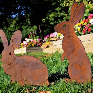 May include: Two rusty metal rabbit garden sculptures. One rabbit is sitting, the other is standing upright. The sculptures are placed in a grassy area with a colorful flower bed in the background. The rabbits have long ears and detailed features.