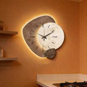 Op de afbeelding: Een moderne wandklok met een uniek ontwerp. De klok heeft een lichtgekleurde cirkelvormige wijzerplaat met zwarte wijzers en een getextureerde, steenachtige buitenrand. De klok wordt verlicht met een warm, zacht licht, wat een stijlvolle en eigentijdse uitstraling creëert.