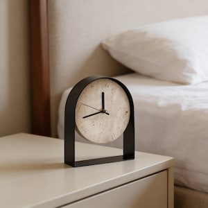 Könnte beinhalten: Eine moderne schwarze Metalluhr mit rundem Zifferblatt. Das Zifferblatt hat ein gesprenkeltes, hellbeige Aussehen. Die Uhr steht auf einem hellbeigen Nachttisch, mit einem Bett und einem Kissen im Hintergrund. Die Zeiger der Uhr sind schwarz.