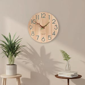 May include: A round wooden wall clock with a light brown finish. The clock features dark green numerals and hands, with a red second hand. The clock face has the text "TIN & TIN" at the bottom. The clock is hanging on a light beige wall.
