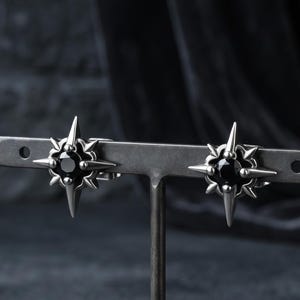 May include: A pair of silver-toned stud earrings featuring a central round black stone set within a four-pointed star design with additional smaller spikes between each point. The earrings are displayed on a horizontal metal bar stand.