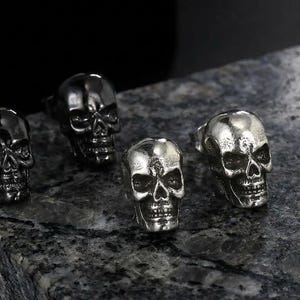 Puede incluir: Cuatro anillos metálicos con forma de calavera dispuestos en grupo. Dos anillos presentan un acabado oscuro y pulido en color gris plomo, mientras que los otros dos muestran un acabado plateado más claro y desgastado con textura.