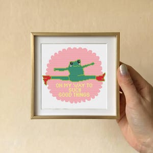 May include: A square gold-toned frame containing a cross-stitch embroidery of a green frog performing a split on a pink scalloped circle background. The text "ON MY WAY TO SUCH GOOD THINGS" is stitched in yellow letters below the frog figure.