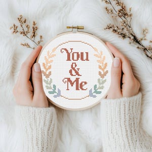Può includere: Un telaio da ricamo a punto croce con le parole "You & Me" in un carattere color ruggine, circondato da una ghirlanda di foglie dai colori pastello. Il telaio è tenuto da mani che indossano un maglione color crema, su uno sfondo bianco soffice.
