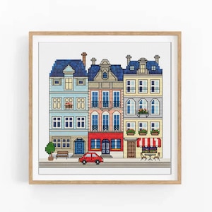 Könnte beinhalten: Kreuzstich-Kunstwerk mit farbenfrohen europäischen Gebäuden. Die Gebäude haben blaue, rosa, rote und gelbe Fassaden mit Fenstern, Balkonen und Markisen. Ein kleines rotes Auto parkt auf der Straße vor den Gebäuden. Das Kunstwerk ist in einem hellen Holzrahmen gerahmt.