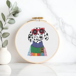 Puede incluir: Un bastidor de bordado de punto de cruz con un perro dálmata con gafas de sol rojas y un suéter colorido. El perro tiene manchas negras sobre un fondo blanco. El bastidor es de madera y tiene un mecanismo de apriete color latón.