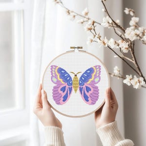 Può includere: Un telaio da ricamo a punto croce con un colorato disegno a farfalla. La farfalla ha ali blu, gialle e viola con motivi intricati. Il ricamo è su sfondo bianco, tenuto in un telaio di legno.