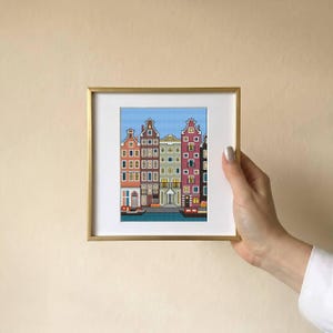 Op de afbeelding: Een ingelijst kruissteekkunstwerk dat een rij kleurrijke grachtenpanden afbeeldt. Het kunstwerk heeft een gouden lijst en een witte passe-partout. De huizen zijn in tinten rood, oranje, groen en paars, met een blauwe lucht op de achtergrond.