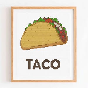 Puede incluir: Una obra de arte en punto de cruz enmarcada que representa un taco detallado. El taco está relleno de ingredientes rojos, verdes y blancos, sobre una concha de color beige claro. La palabra "TACO" está bordada en letras marrones debajo.