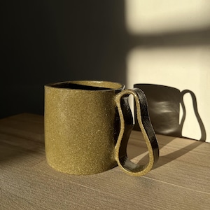 Peut inclure: Une tasse en céramique faite à la main, de couleur vert olive moucheté, avec une anse originale en forme de boucle. L'intérieur est sombre. La tasse est posée sur une surface en bois, avec son ombre projetée sur un mur.